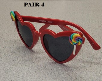 May include: Red heart-shaped sunglasses with dark lenses and a rainbow lollipop accent on each side. The sunglasses are branded "FOSTER GRANT" and the text "PAIR 4" is visible in the upper left corner.