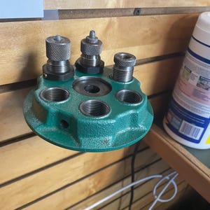 May include: A reloading die set with a green base and three silver dies. The dies are attached to the base with threaded connections. The base has several threaded holes for the dies and a central opening.