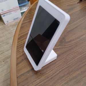 May include: A white tablet with a black screen is propped up on a white stand. The tablet is rectangular with rounded corners. The stand is a simple, rectangular shape, designed to hold the tablet upright.