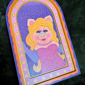 May include: A framed artwork featuring Miss Piggy from The Muppets. The artwork has a purple background with a glittery effect, and Miss Piggy is wearing a pink dress and a pearl necklace. The frame is multi-layered with orange, pink, and blue.