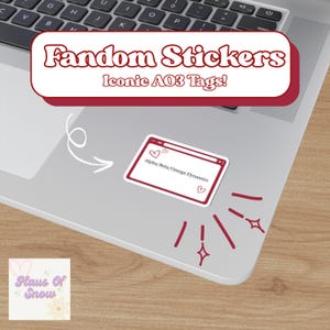 Pode incluir: Um laptop com um adesivo que diz "Fandom Stickers Iconic AO3 Tags!" e um adesivo com o texto "Alpha/Beta/Omega Dynamics" e corações. O laptop está em uma superfície de madeira.