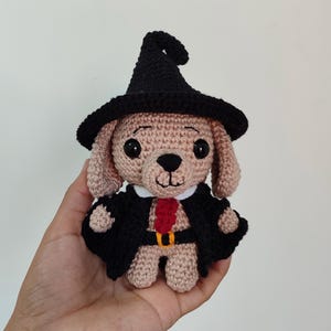 May include: A crocheted dog wearing a black witch hat, a black robe, and a red tie. The dog has brown fur and black eyes.