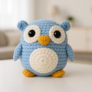 May include: A handmade, blue crochet owl with large black eyes, a yellow beak, and yellow feet. The owl has a white belly and is sitting on a white surface. The owl is a decorative item.
