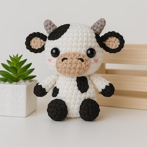 Può includere: Un peluche di mucca fatto a mano all'uncinetto con corpo bianco, macchie nere e zoccoli neri. La mucca ha un muso marrone, guance rosa e grandi occhi neri. Le orecchie sono nere e marroni. Il peluche è su una superficie bianca.