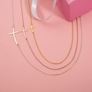 May include: Three delicate gold chains with a cross pendant on each. The chains are layered on a pink background.