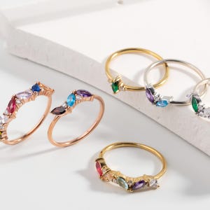 May include: Five rings with colorful gemstones set in a delicate marquise shape. The rings are made of gold and silver and are arranged on a white surface.