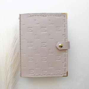 May include: A light taupe leather wallet with the embossed letters "L E L E L E" repeated across the front. The wallet has a snap closure with a gold-colored button and gold corner accents. The stitching is visible around the edges.