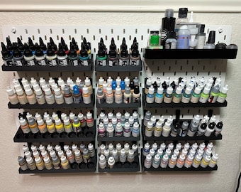 May include: A white pegboard wall with multiple rows of small bottles of paint organized in black plastic holders. The bottles are arranged in rows of different colors, including black, white, brown, blue, green, and red.