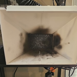 Può includere: Una cabina di verniciatura in plastica bianca con una rete a maglie nere e un tubo nero attaccato al lato. L'interno della cabina è sporco con schizzi di vernice nera.