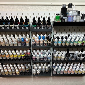 May include: A white pegboard wall with multiple rows of small bottles of paint organized in black plastic holders. The bottles are arranged in rows of different colors, including black, white, brown, blue, green, and red.