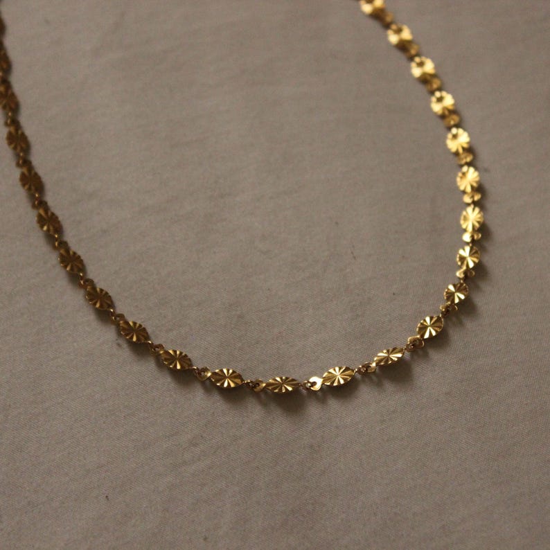 Puede incluir: Un delicado collar dorado con un patr&oacute;n repetitivo de peque&ntilde;os eslabones en forma de flor. La cadena se muestra sobre un fondo neutro, destacando el intrincado dise&ntilde;o y el c&aacute;lido tono met&aacute;lico de la joya.
