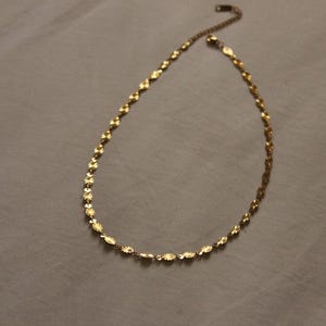 Puede incluir: Un delicado collar dorado con una cadena y peque&ntilde;os colgantes con forma de flor. El collar se muestra sobre una superficie de color neutro. La cadena es ajustable y tiene un cierre de langosta.