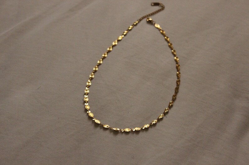 Puede incluir: Un delicado collar dorado con una cadena y peque&ntilde;os colgantes con forma de flor. El collar se muestra sobre una superficie de color neutro. La cadena es ajustable y tiene un cierre de langosta.