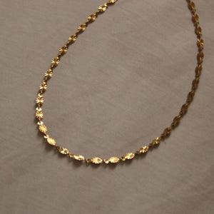 Puede incluir: Un delicado collar dorado con un patr&oacute;n repetitivo de peque&ntilde;os eslabones ovalados y en forma de estrella texturizados. El collar se muestra sobre una superficie gris neutra, resaltando su intrincado dise&ntilde;o y c&aacute;lido tono met&aacute;lico. Adecuado para uso diario o especial.