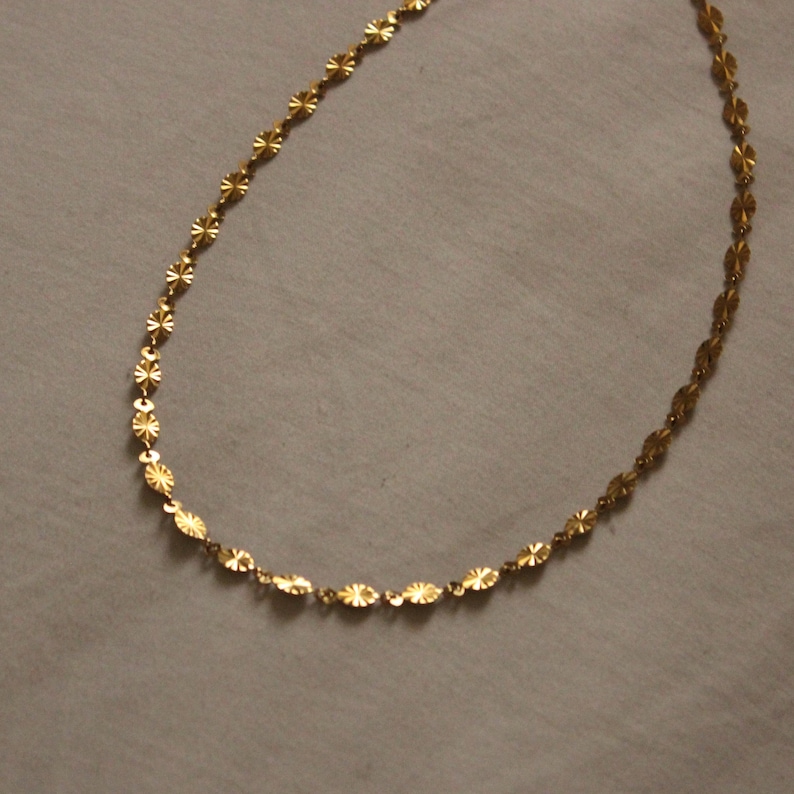 Puede incluir: Un delicado collar dorado con un patr&oacute;n repetitivo de peque&ntilde;os eslabones ovalados y en forma de estrella texturizados. El collar se muestra sobre una superficie gris neutra, resaltando su intrincado dise&ntilde;o y c&aacute;lido tono met&aacute;lico. Adecuado para uso diario o especial.