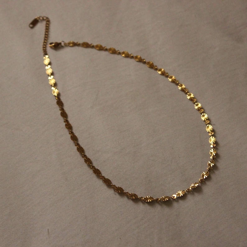 Puede incluir: Un delicado collar dorado con un dise&ntilde;o floral. El collar presenta peque&ntilde;os dijes en forma de flor unidos para formar una cadena. El collar se muestra sobre un fondo neutro. Mide aproximadamente 40 cm.