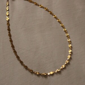 Puede incluir: Un delicado collar de color dorado con un dise&ntilde;o floral repetitivo. El collar est&aacute; compuesto por peque&ntilde;os eslabones planos en forma de flor, conectados para formar una cadena. La cadena se muestra sobre un fondo neutro.