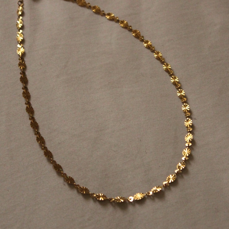 Puede incluir: Un delicado collar de color dorado con un dise&ntilde;o floral repetitivo. El collar est&aacute; compuesto por peque&ntilde;os eslabones planos en forma de flor, conectados para formar una cadena. La cadena se muestra sobre un fondo neutro.