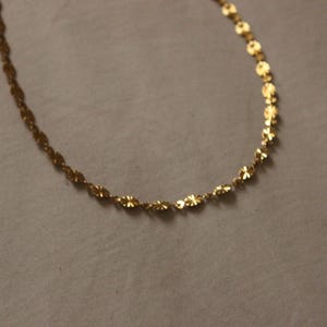 Puede incluir: Un delicado collar de color dorado con un dise&ntilde;o floral. La cadena est&aacute; compuesta por peque&ntilde;os eslabones interconectados en forma de flor. El collar se muestra sobre un fondo neutro, resaltando sus intrincados detalles y su c&aacute;lido tono.