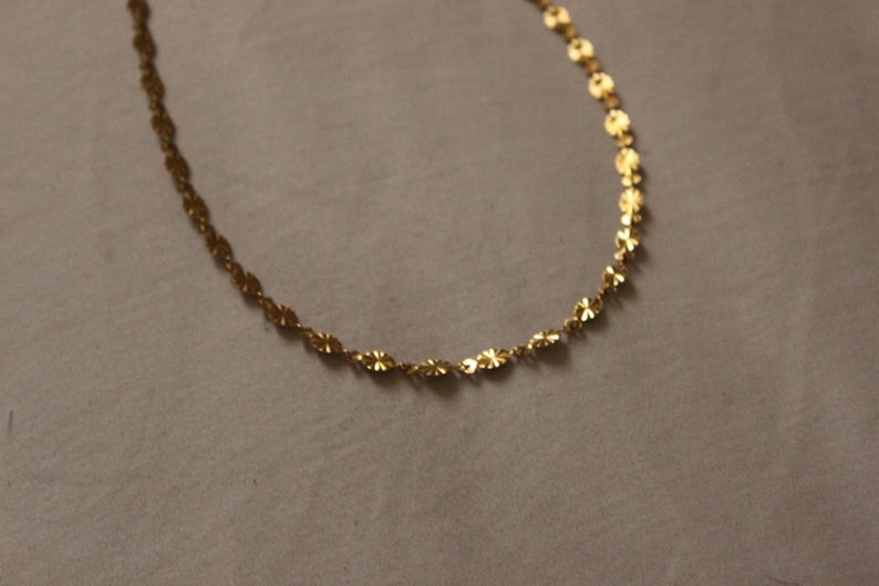 Puede incluir: Un delicado collar de color dorado con un dise&ntilde;o floral. La cadena est&aacute; compuesta por peque&ntilde;os eslabones interconectados en forma de flor. El collar se muestra sobre un fondo neutro, resaltando sus intrincados detalles y su c&aacute;lido tono.