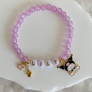 Pode incluir: Um bracelete de contas roxas com um fecho de ouro que diz "LILY" e um fecho de personagem de desenho animado preto e branco. O bracelete tamb&eacute;m tem um fecho de estrela dourada e um fecho de cora&ccedil;&atilde;o.