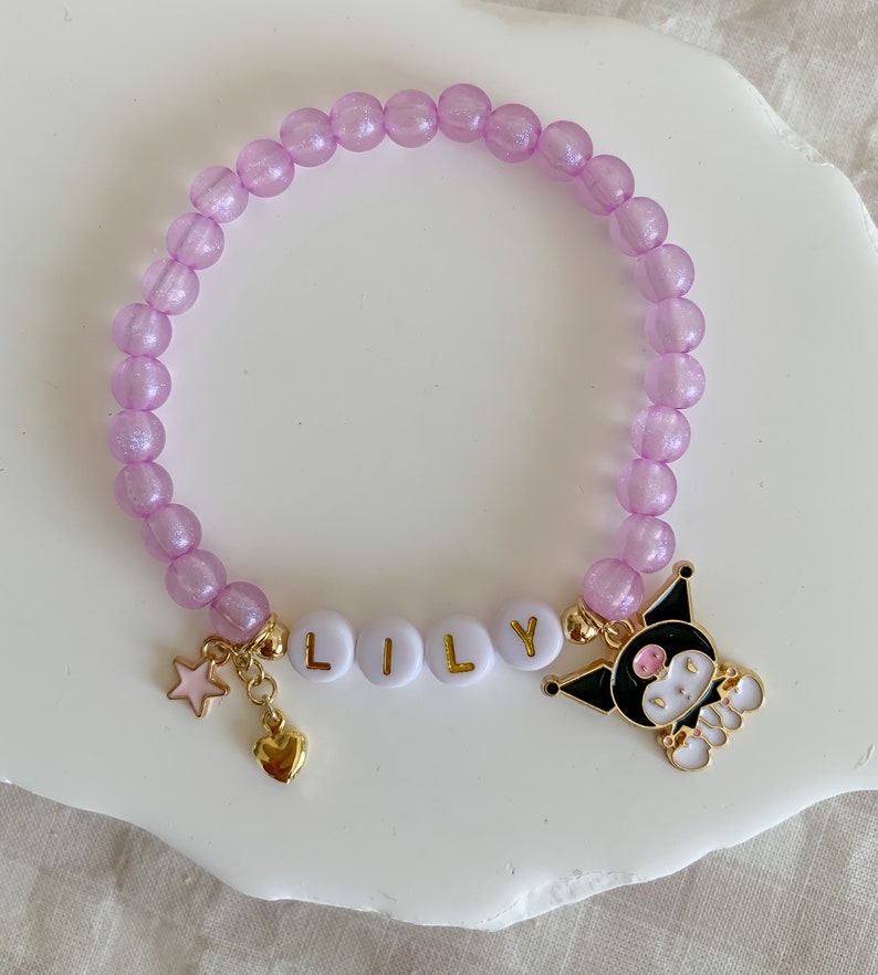 Pode incluir: Um bracelete de contas roxas com um fecho de ouro que diz "LILY" e um fecho de personagem de desenho animado preto e branco. O bracelete tamb&eacute;m tem um fecho de estrela dourada e um fecho de cora&ccedil;&atilde;o.