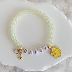 Pode incluir: Um bracelete de contas amarelo claro com um amuleto de ouro de um c&atilde;o de desenho animado e um amuleto de ouro com o nome "DAISY" escrito em letras brancas.