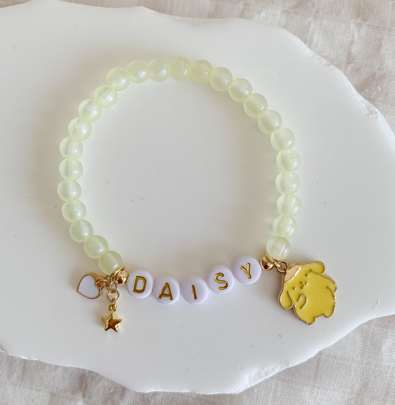 Pode incluir: Um bracelete de contas amarelo claro com um amuleto de ouro de um c&atilde;o de desenho animado e um amuleto de ouro com o nome "DAISY" escrito em letras brancas.