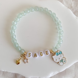 Pode incluir: Um bracelete de contas verde claro com pingentes dourados. Os pingentes incluem um cora&ccedil;&atilde;o, uma estrela, o nome "SAGE" escrito em letras douradas e um pingente Hello Kitty.