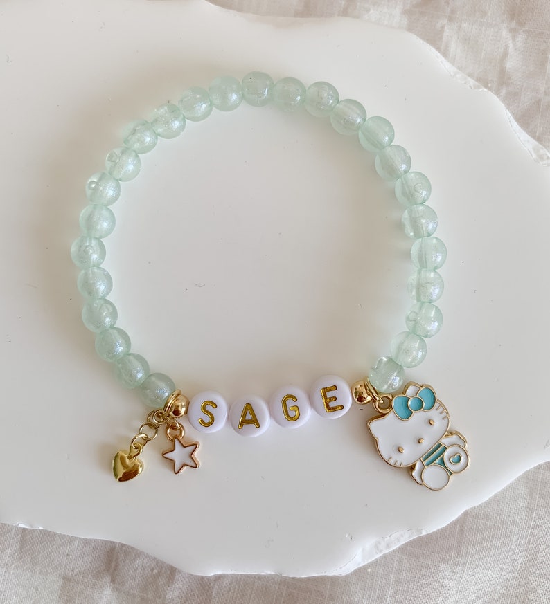 Pode incluir: Um bracelete de contas verde claro com pingentes dourados. Os pingentes incluem um cora&ccedil;&atilde;o, uma estrela, o nome "SAGE" escrito em letras douradas e um pingente Hello Kitty.