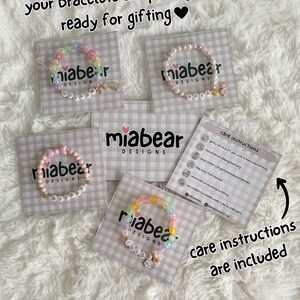 Pode incluir: Quatro pulseiras de contas coloridas com nomes personalizados em cart&otilde;es brancos. Cada pulseira tem um amuleto. Os cart&otilde;es t&ecirc;m um padr&atilde;o xadrez rosa e branco. Tamb&eacute;m est&aacute; inclu&iacute;do um cart&atilde;o com instru&ccedil;&otilde;es de cuidados.