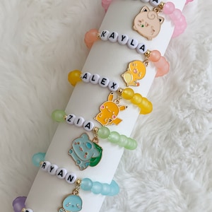 May include: Six colourful beaded bracelets with charms featuring cartoon characters. Each bracelet has a name spelled out in white beads: Kym, Lana, Kayla, Alex, Zia, Ryan, and Eli.