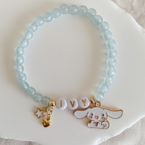 Pode incluir: Um bracelete de contas azul claro com um fecho dourado com um c&atilde;o de desenho animado branco e o nome "IVY" escrito em letras douradas.