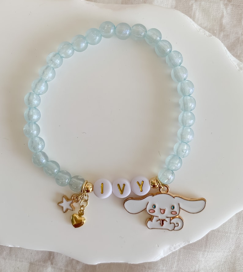 Pode incluir: Um bracelete de contas azul claro com um fecho dourado com um c&atilde;o de desenho animado branco e o nome "IVY" escrito em letras douradas.