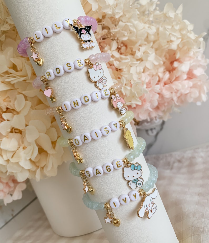 Pode incluir: Cinco pulseiras de contas coloridas com pingentes. Cada pulseira tem um nome diferente escrito em letras douradas: Lily, Rose, Indie, Daisy e Sage. Os pingentes incluem uma estrela, um cora&ccedil;&atilde;o e um personagem de desenho animado.