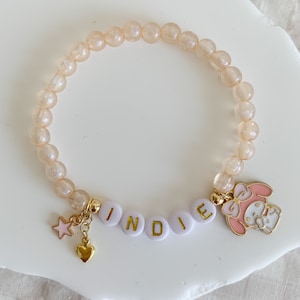 Pode incluir: Um bracelete rosa claro e dourado com um amuleto que diz "INDIE" e um amuleto de personagem de desenho animado rosa. O bracelete &eacute; feito de pequenas contas redondas transl&uacute;cidas.