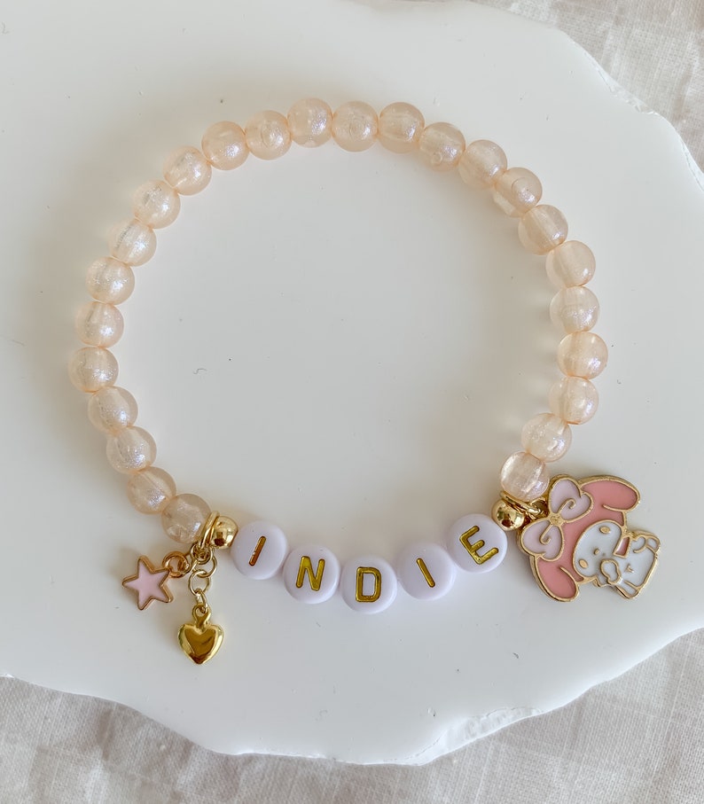 Pode incluir: Um bracelete rosa claro e dourado com um amuleto que diz "INDIE" e um amuleto de personagem de desenho animado rosa. O bracelete &eacute; feito de pequenas contas redondas transl&uacute;cidas.