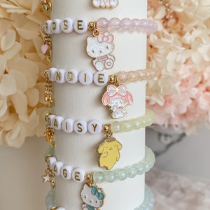 Pode incluir: Seis pulseiras de contas coloridas com pingentes que representam personagens de desenhos animados populares. Cada pulseira tem um nome escrito em contas brancas: Lily, Rose, Indie, Daisy, Sage e Ivy.