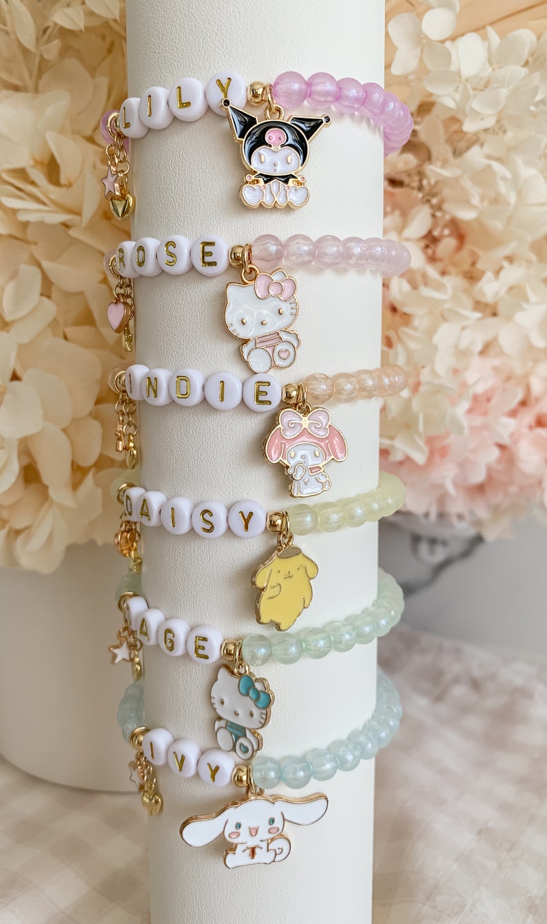Pode incluir: Seis pulseiras de contas coloridas com pingentes que representam personagens de desenhos animados populares. Cada pulseira tem um nome escrito em contas brancas: Lily, Rose, Indie, Daisy, Sage e Ivy.