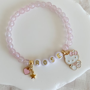 Pode incluir: Um bracelete de contas cor-de-rosa com um amuleto branco que diz "ROSE" e um amuleto dourado de um gato de desenho animado com um la&ccedil;o rosa. O bracelete tamb&eacute;m tem um amuleto dourado de cora&ccedil;&atilde;o e estrela.