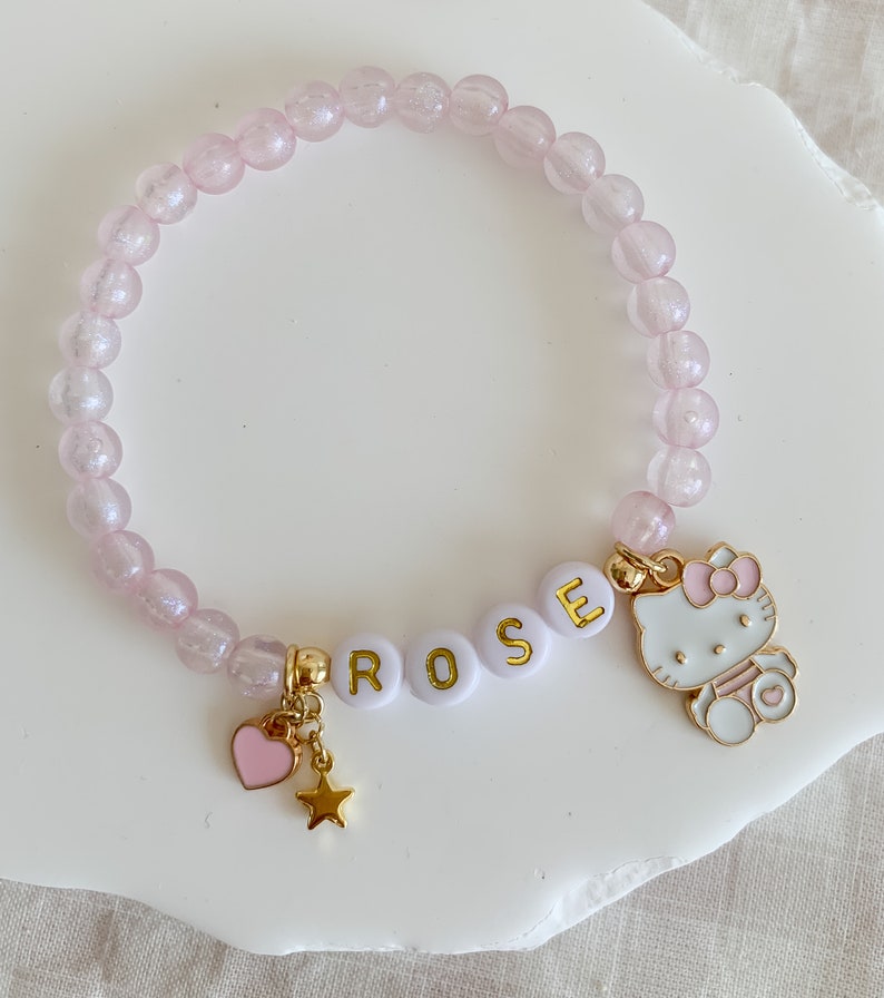 Pode incluir: Um bracelete de contas cor-de-rosa com um amuleto branco que diz "ROSE" e um amuleto dourado de um gato de desenho animado com um la&ccedil;o rosa. O bracelete tamb&eacute;m tem um amuleto dourado de cora&ccedil;&atilde;o e estrela.