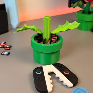 May include: A green 3D-printed planter shaped like a pipe, holding video game cartridges. A black and white controller is in front of the planter. The planter has green leaves and a central stem. The controller has blue and orange buttons.