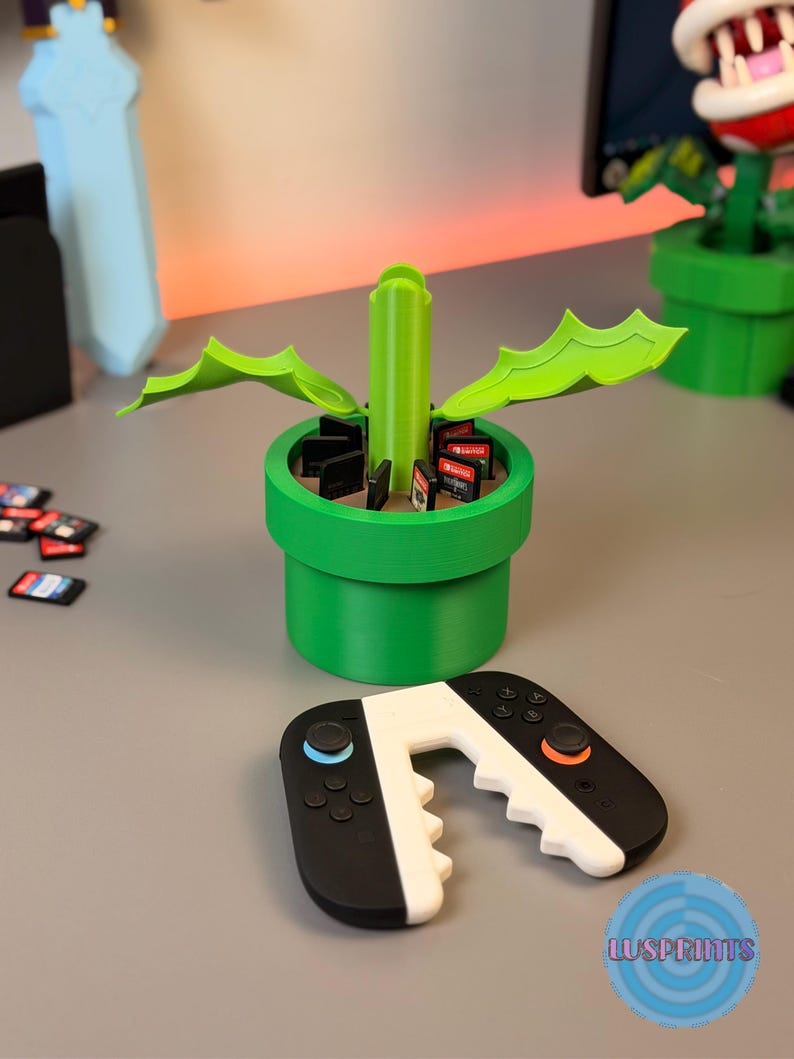 May include: A green 3D-printed planter shaped like a pipe, holding video game cartridges. A black and white controller is in front of the planter. The planter has green leaves and a central stem. The controller has blue and orange buttons.