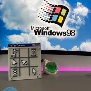 May include: A 3D-printed tic-tac-toe game with hourglass and arrow icons, next to a small, white container with green contents. The background features a computer screen displaying the Microsoft Windows 98 logo against a blue sky.