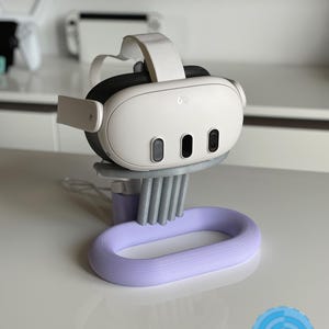 May include: A white and black VR headset with a white strap sits on a gray and lavender stand. The stand has a lavender oval base and a series of gray vertical supports. The headset has three black sensors and the Meta logo.