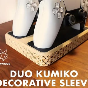 May include: A beige decorative sleeve for game controllers, featuring a geometric pattern. The sleeve holds two white controllers. The text "DUO KUMIKO DECORATIVE SLEEVE" is displayed below the sleeve.