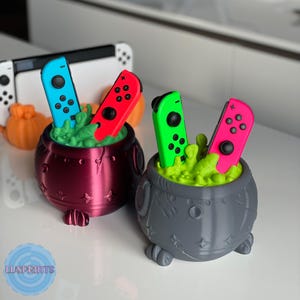 May include: Two cauldron-shaped Nintendo Switch controller holders. One is dark red, the other gray. Each holds two Joy-Con controllers, one blue and red, the other green and pink, set in green, bubbling, stylized liquid. A Switch console is in the background.
