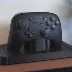 May include: A black Nintendo Switch Pro Controller resting on a matching charging dock. The controller features a D-pad, analog sticks, and action buttons. The dock has blue and red accents. The controller and dock are on a wooden surface.