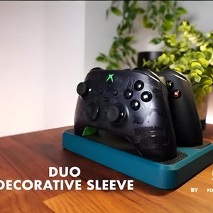 May include: Black and clear Xbox controllers on a teal base. The controllers have green and red accents. The text "DUO DECORATIVE SLEEVE" is displayed on the base. The background includes a plant and a wooden surface.
