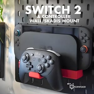 May include: Black wall mount for a Switch 2 and controller. The mount is black with red accents and holds the Switch and controller. The text "SWITCH 2 & CONTROLLER WALL/SKADIS MOUNT" is displayed on the top of the mount.